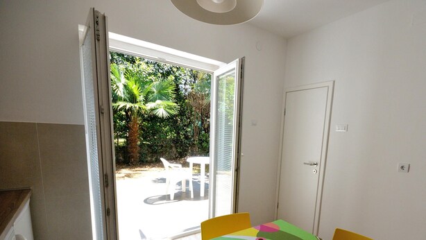 Ein Zimmer mit einem Tisch, der mit einer bunten Tischdecke bedeckt ist, einer Tür und Blick auf einen Garten durch eine geöffnete Tür.
