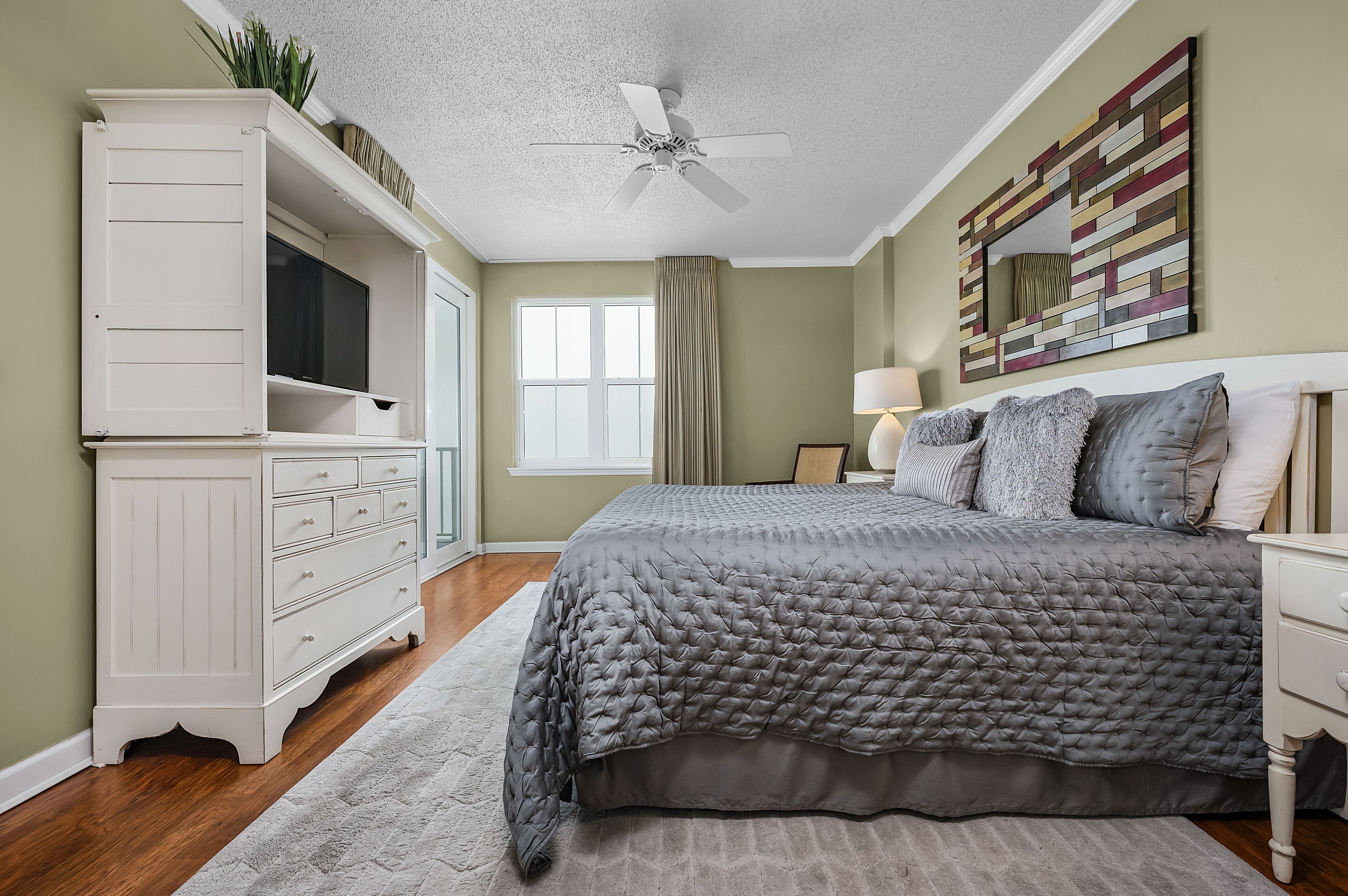 A bedroom with a bed, a dresser, a television, a ceiling fan, a window, and a painting on the wall.