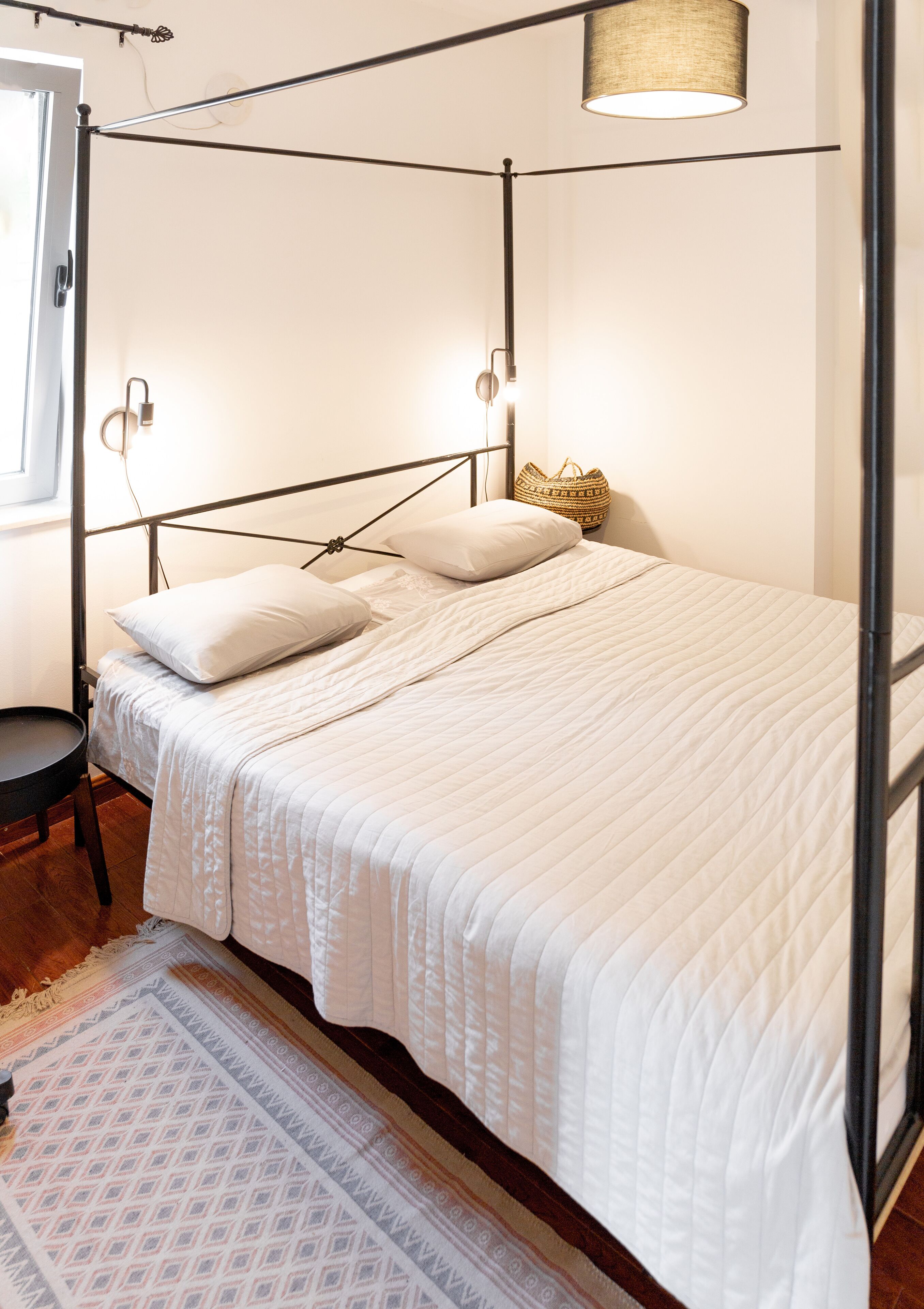 Una cama bien hecha con colcha blanca, cabecera de metal negro y lámpara de noche.