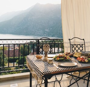 Un balcon avec une table dressée pour un repas, offrant une vue sur un paysage montagneux et une étendue d’eau.