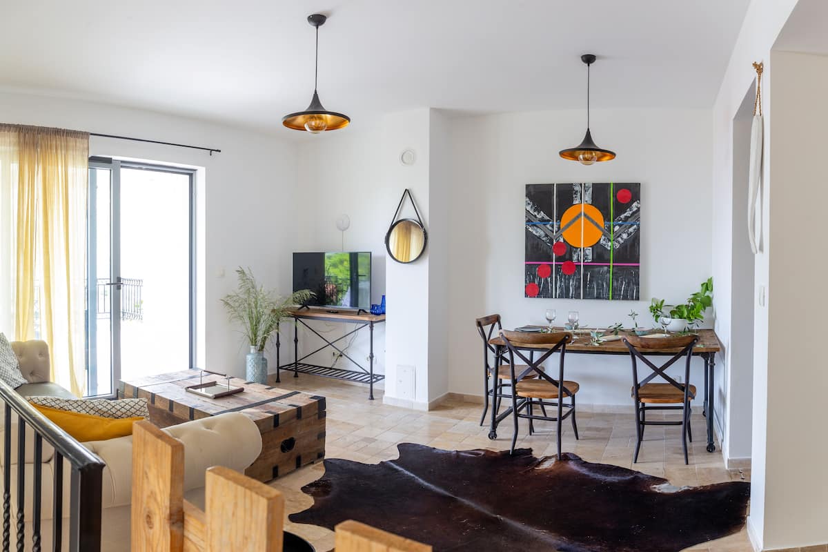 Una sala de estar moderna con un sofá, una mesa de centro de madera y un comedor con mesa y sillas. Hay un ventanal con cortinas, una lámpara colgante y un cuadro en la pared.