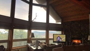 Ein gemütliches Wohnzimmer mit großen Fenstern, einem steinernen Kamin und einem Fernseher, der ein Sportereignis zeigt.