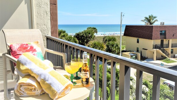 Un balcon avec vue sur la plage, une table avec une serviette, une boisson et une bouteille de Corona Extra.