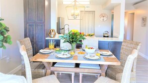 Un coin repas avec une table en bois dressée pour quatre personnes, entourée de chaises en osier. Un lustre est suspendu au plafond et une horloge est visible au mur.