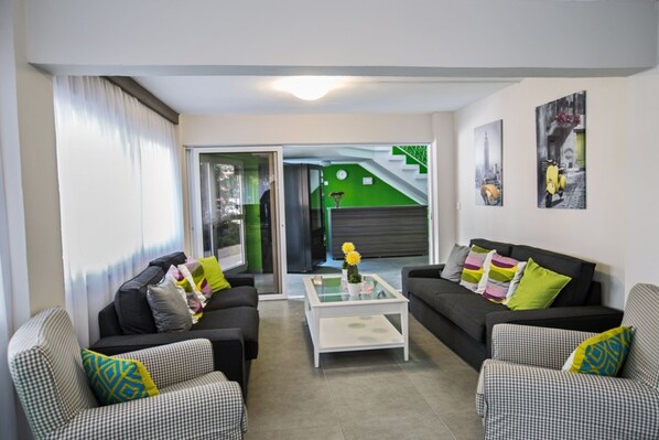 A living room with two black sofas, a white coffee table, and patterned armchairs. There is a glass door leading to a green area with a staircase.