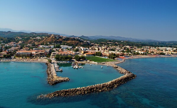 Un pueblo costero con puerto deportivo, playa de arena y montañas al fondo.