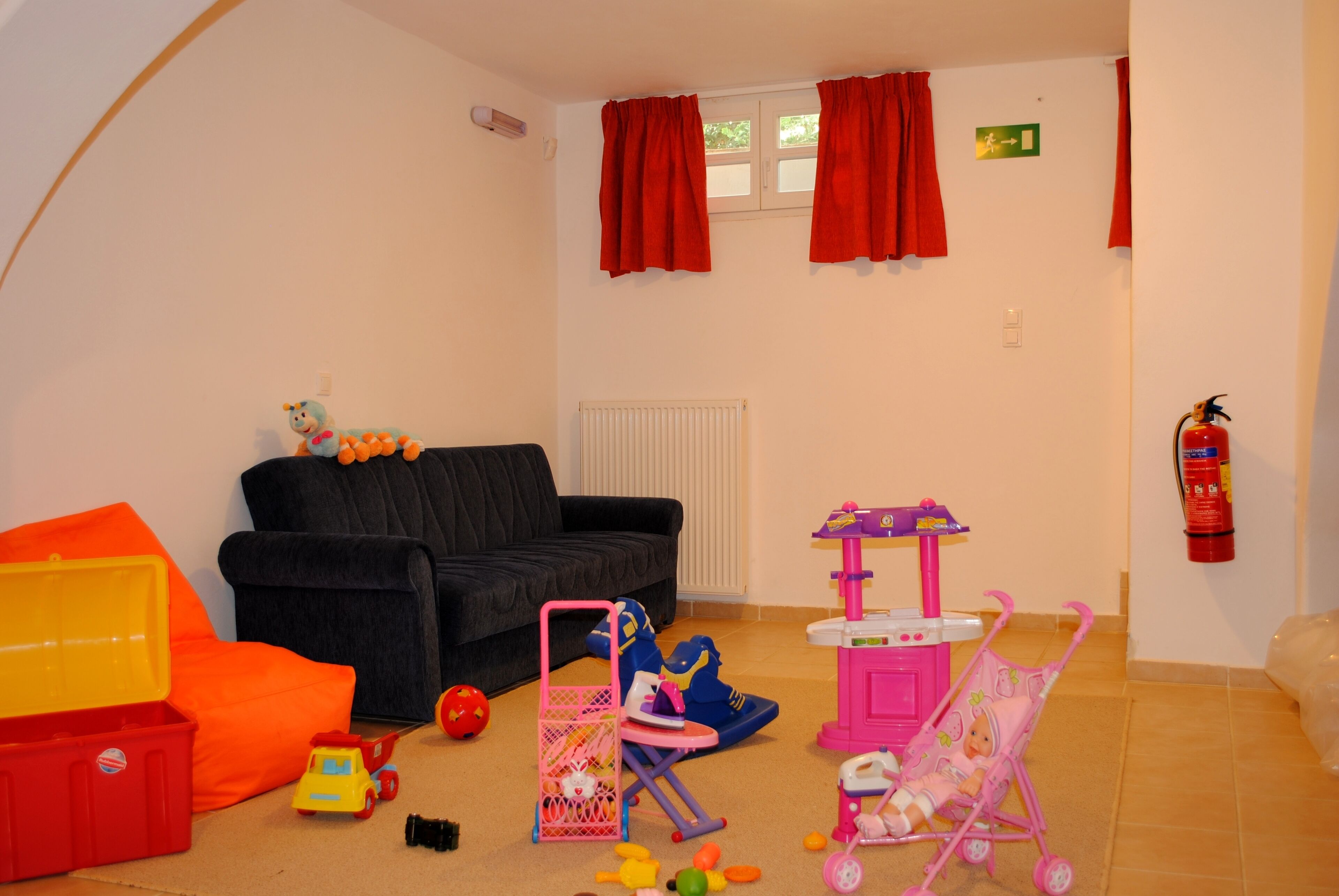 Un salon avec un canapé noir, un fauteuil rouge et une petite table avec des jouets. Il y a une fenêtre avec des rideaux rouges et un extincteur accroché au mur.