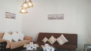 A living room with a brown sofa, a beige sofa, and a coffee table with a floral tablecloth. There are two wall-mounted light fixtures and two paintings on the walls.