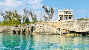 A coastal house with a balcony overlooking turquoise water, situated on a rocky shoreline with sparse vegetation. - High-end New Home - Large PRIVATE Pool, Snorkeling, Cliff Jumping, Kayaks & SUP (Eleuthera)