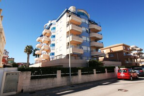 A multi-story residential building with balconies, a street in front, and a palm tree. - Apartment with pool and sea views. (Oliva)