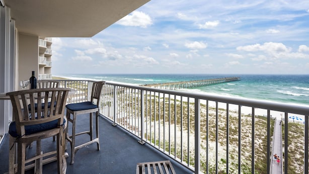 A balcony with two wooden chairs and a table, overlooking a beach with a pier. - Summerwind Resort 602 Center * Beach Chair Set Included * Salt Water Vacations (Navarre)