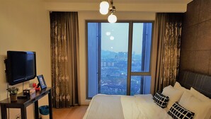 Ein Hotelzimmer mit einem großen Bett, einem Fernseher, einem Schreibtisch mit Pflanze und Blick auf eine Stadt durch ein großes Fenster.