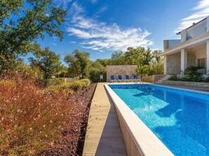 A swimming pool with a wooden deck, surrounded by greenery and a stone wall.