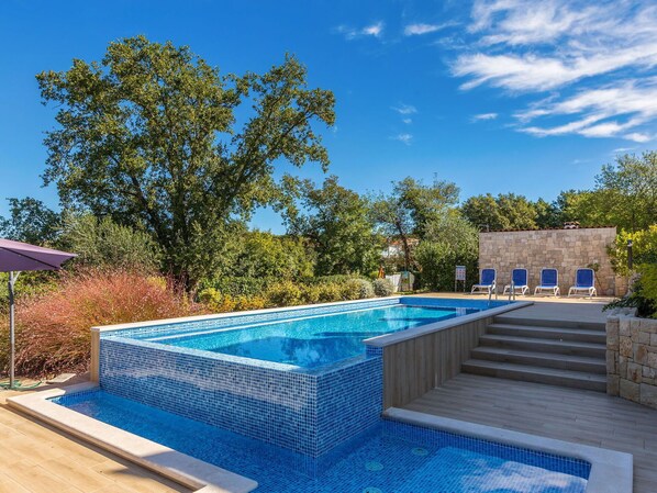 A rectangular swimming pool with blue mosaic tiles, surrounded by a wooden deck and stone walls.