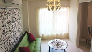 A living room with a green sofa, a glass coffee table, and a decorative ceiling light. - N ° 5  CONDOMINIUM: First floor Corner Apartment ;  Safe Residential Area. (Osaka)