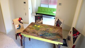 A dining area with a table covered in abstract art, two chairs with colorful patterns, a green sofa in the background, and a chandelier. - N ° 5  CONDOMINIUM: First floor Corner Apartment ;  Safe Residential Area. (Osaka)