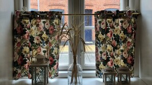 A window with two floral curtains, a clear view of a brick building, and a vase with dried plants on the windowsill. - Cozy town house in the center of Assens, close to the harbor. (Assens)
