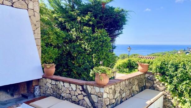 A stone bench with cushions, potted plants, and a view of the sea. - Villenpark Playa Arena House Ocean (Lloret de Mar)