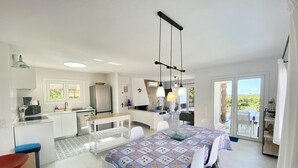 A modern kitchen with white cabinets, a dining area with a patterned tablecloth, and a view of the sea through the sliding glass doors. - Villenpark Playa Arena House Ocean (Lloret de Mar)