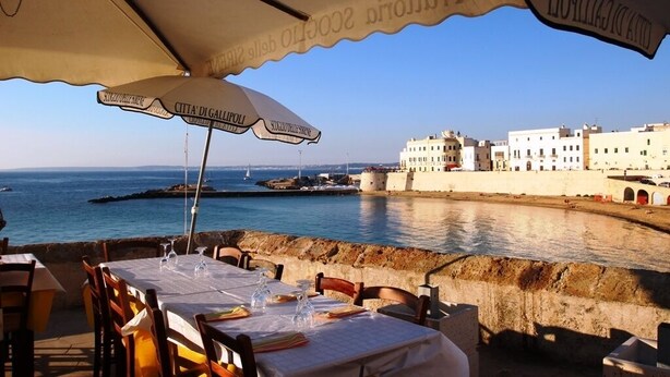 An outdoor dining area with tables set under umbrellas, overlooking a coastal cityscape with historic buildings and a clear view of the sea. - Villa with large private pool, stunning views, fantastic for cycling/ walking (Martina Franca)