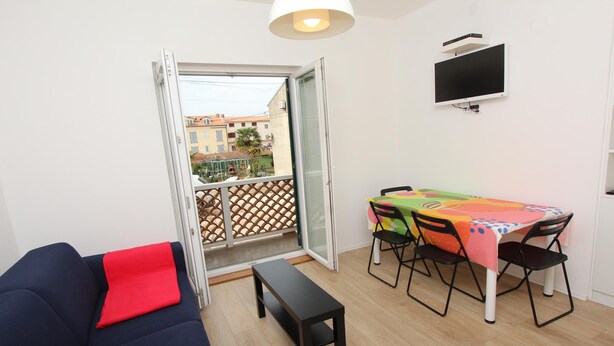 Un petit salon comprenant un canapé, une table avec des chaises, un téléviseur fixé au mur et une porte-fenêtre coulissante donnant sur un balcon.