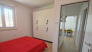 Une chambre avec un lit rouge, une armoire blanche et une porte donnant sur un coin repas équipé de chaises et d’une table.