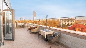 A rooftop terrace with outdoor seating, a glass door entrance, and a view of city buildings. - Praga Hotel (Tashkent)
