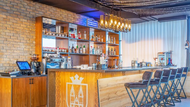 A bar area with wooden shelves stocked with bottles, a coffee machine, and a laptop on the counter. There are foldable chairs and a wooden counter with a crest emblem. - Praga Hotel (Tashkent)