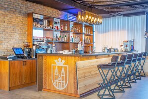 A bar area with wooden shelves stocked with bottles, a coffee machine, and a laptop on the counter. There are foldable chairs and a wooden counter with a crest emblem.