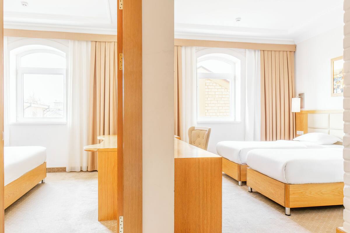 A hotel room with a bed, a desk, and a chair. There is a window with curtains and a view of buildings outside.