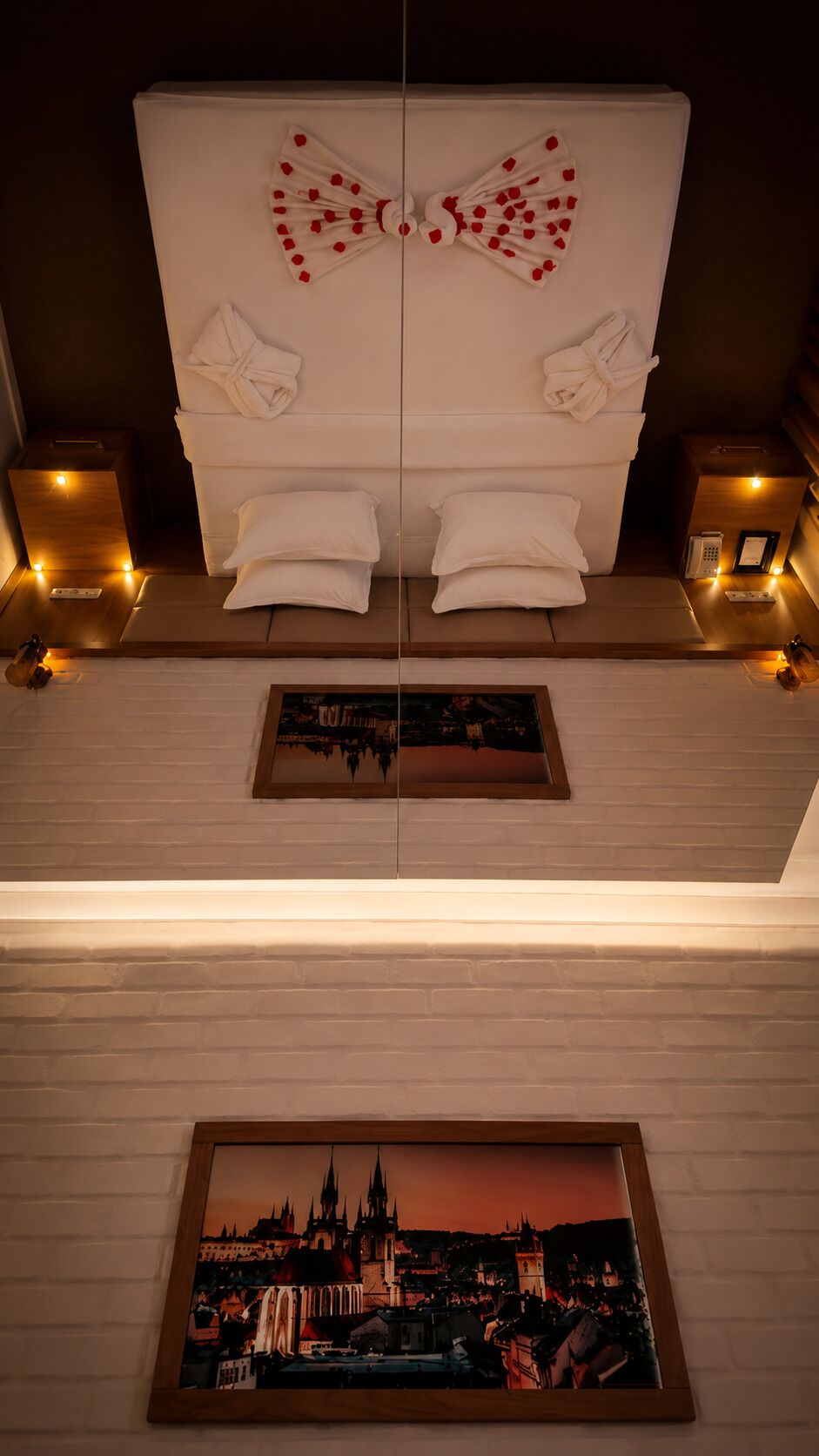 A hotel room with a mirrored ceiling, two white beds with towels, bedside lamps, and a framed cityscape painting on the wall.