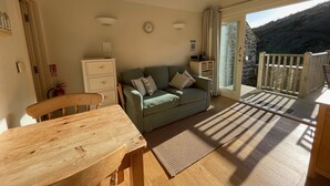 En hyggelig stue med et træbord og stole, en grøn sofa med puder, en hvid kommode og en lille lampe. Der er et vindue med gardiner og udsigt til et bjerglandskab.
