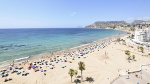 Una platja amb ombrelles i palmeres, cel blau i edificis al fons.
