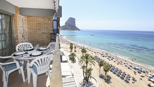Un balcó amb una taula preparada per dinar, amb vista a una platja amb ombrel·les i palmeres.