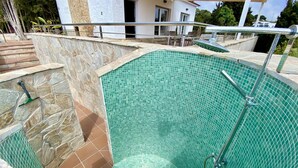 A swimming pool with a stone and tiled border, a metal ladder, and a covered seating area in the background. - Villenpark Playa Arena House Torre Isabel (Lloret de Mar)