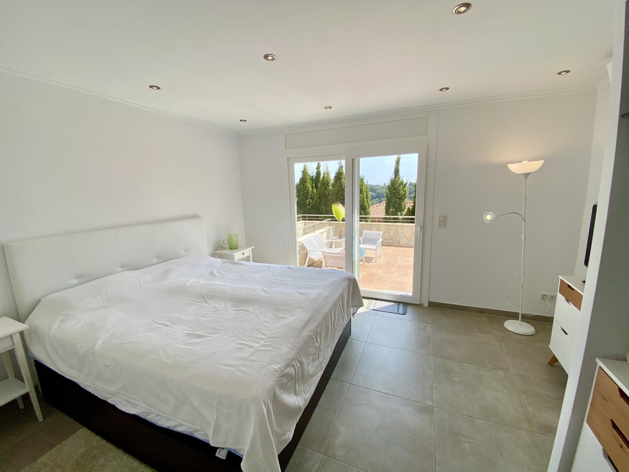 Ein Schlafzimmer mit einem großen Bett, einem Nachttisch, einer Stehlampe und einer Schiebetür zu einem Balkon mit Gartenmöbeln.