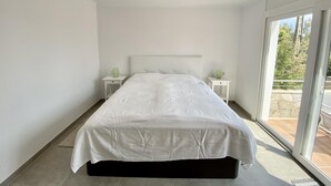 A modern bedroom with a large bed, two bedside tables, and a view of a garden through a sliding glass door. - Villenpark Playa Arena House Torre Isabel (Lloret de Mar)