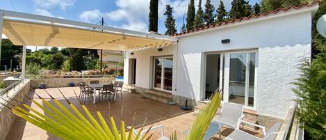Un patio cubierto con mesa y sillas, una piscina y un edificio blanco con amplias ventanas.