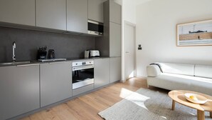 Eine moderne Küche mit grauen Schränken, einem eingebauten Backofen und einer Kaffeemaschine. Es gibt einen Holzboden, ein weißes Sofa und einen kleinen Holztisch mit einem Teller.