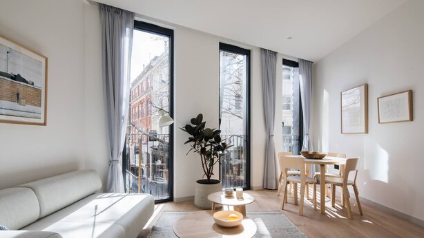 Ein modernes Wohnzimmer mit einer Couch, einem Esstisch mit Stühlen, einem Couchtisch und großen Fenstern mit Vorhängen.