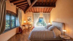 A bedroom with a large bed, a desk, and a chair. There is a window with curtains, a lamp, and a painting on the wall.