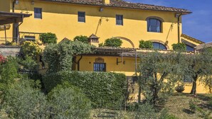 A yellow villa with multiple windows, a balcony, and a garden with olive trees.