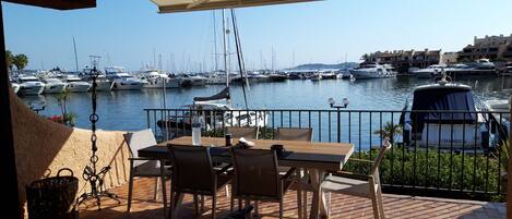 A terrace with a view of a marina, featuring a table and chairs, a wicker basket, and a covered area with a canopy.