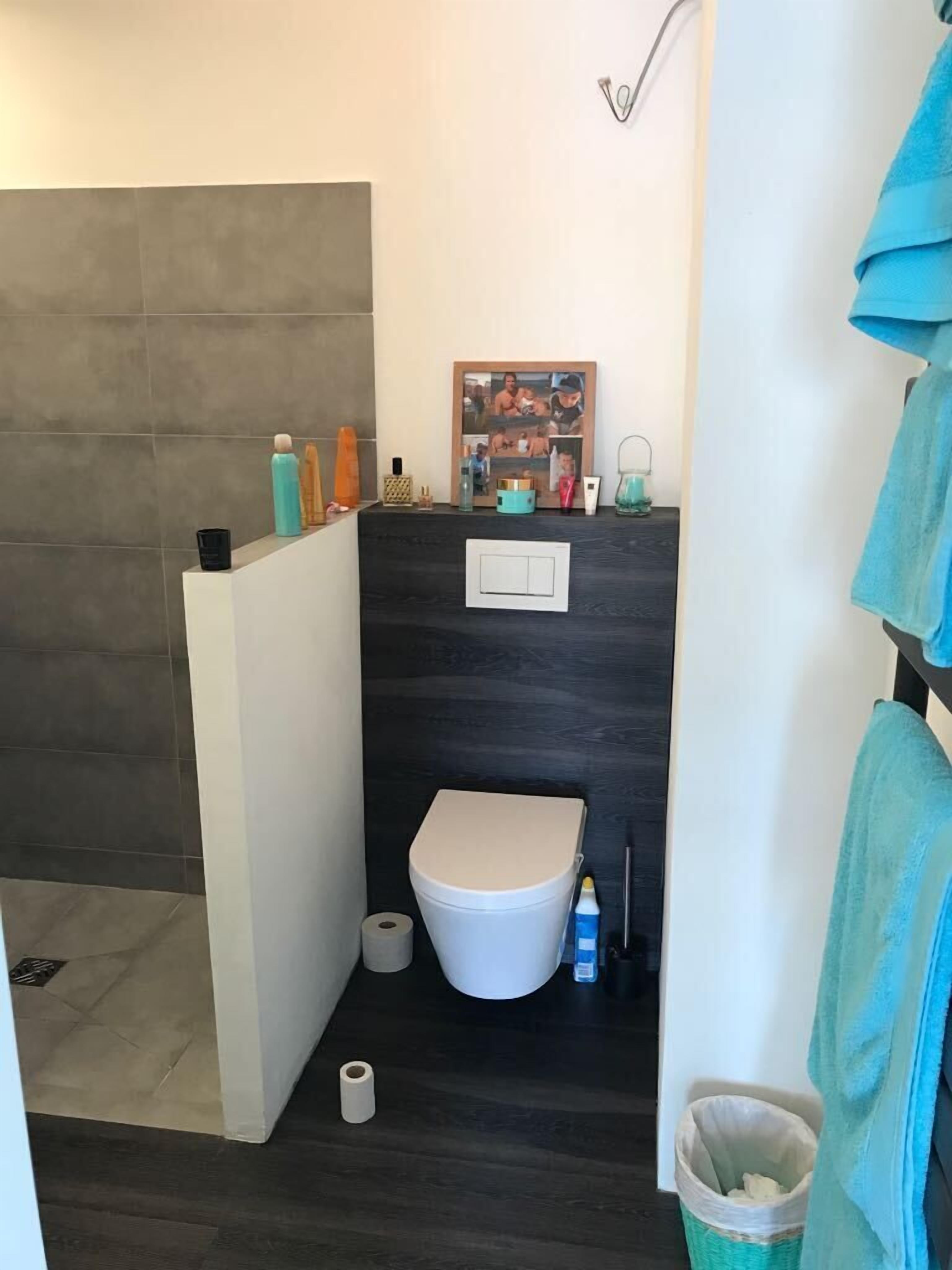 A compact bathroom with a toilet, a small sink, and a shower area. There are toiletries on the counter, and a picture frame is mounted on the wall.