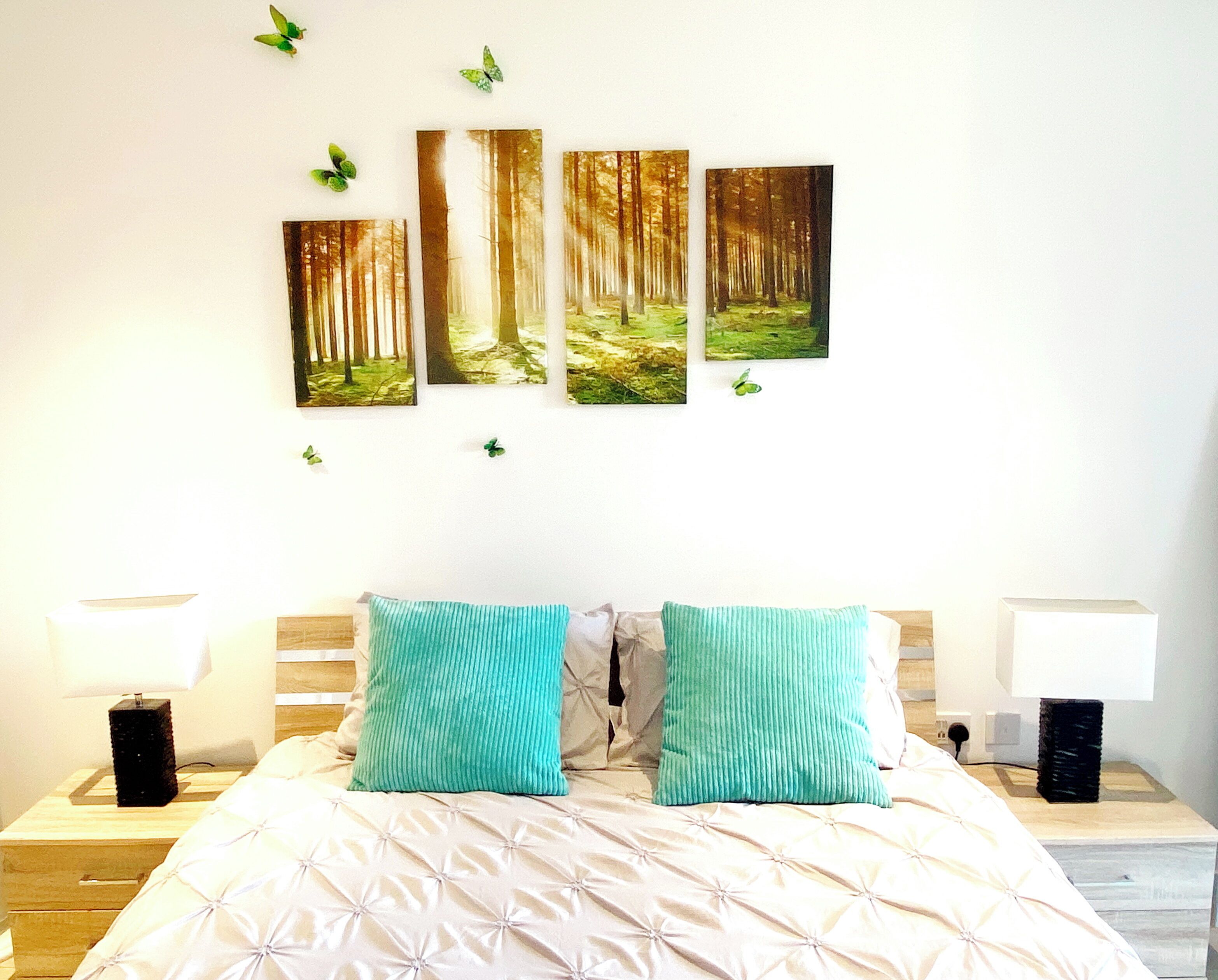 Een slaapkamer met een bed, twee nachtkastjes, twee wandlampen en vier afbeeldingen van een bos in lijsten.
