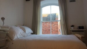 A bedroom with a large bed, a window with curtains, a radiator, and a small plant on a shelf.