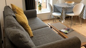 A living room with a grey sofa, a small table, and a dining area with a patterned tablecloth.