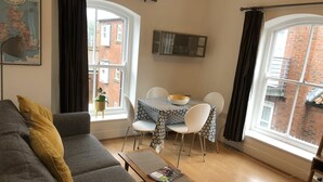 A living room with a grey sofa, a wooden coffee table, and a dining table with white chairs. There are two windows with curtains, a map on the wall, and a small plant on the windowsill.