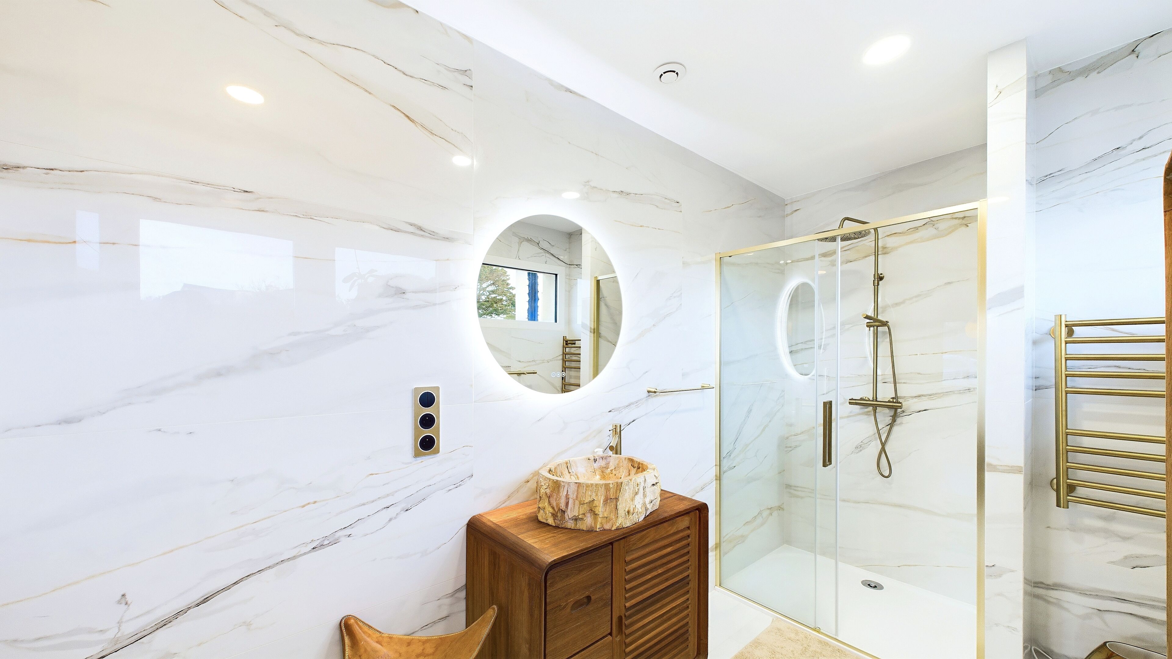 A modern bathroom with a marble wall, a wooden vanity, a glass shower enclosure, and a circular mirror.
