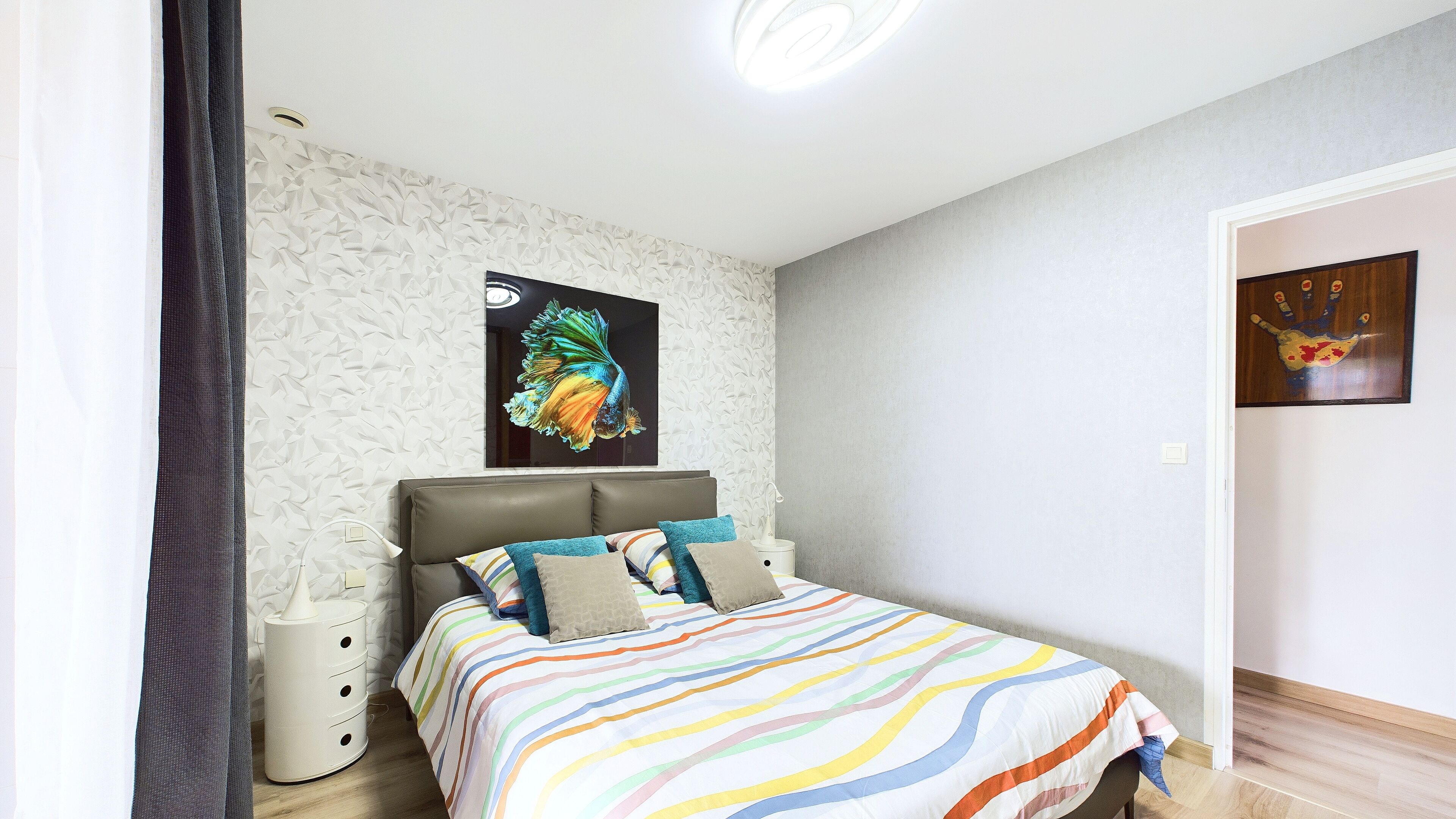 Une chambre à coucher avec une couvre-lit à rayures, une tête de lit, une table de chevet, une œuvre d’art accrochée au mur et une porte ornée d’un motif représentant une empreinte de main.